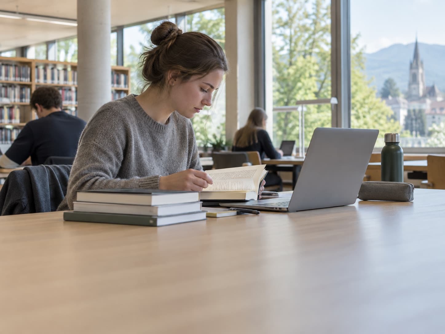 Une jeune femme lit à une table de bibliothèque, un ordinateur portable ouvert et une pile de livres devant elle.