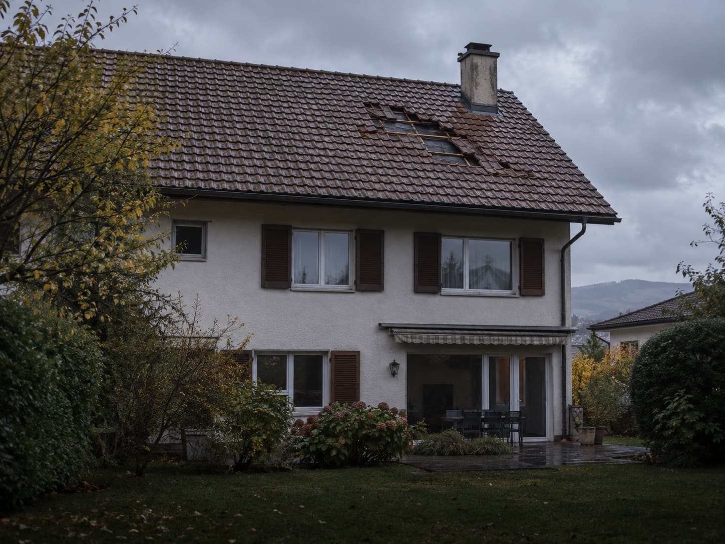 Une maison vue depuis le jardin sous un ciel couvert, avec plusieurs tuiles déplacées ou manquantes sur le toit.