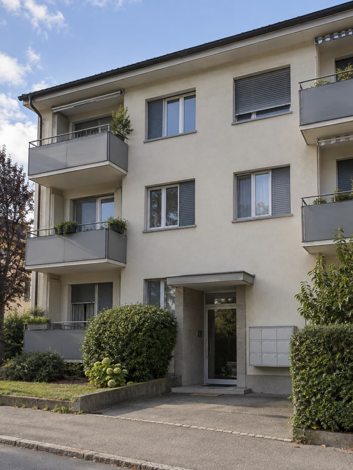 La façade claire d’un petit immeuble avec des balcons et des stores gris, une entrée vitrée et des boîtes aux lettres, vue depuis le trottoir.